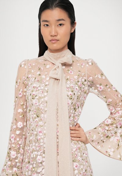 Γυναίκα με μακριά σκούρα μαλλιά φορεί ένα διάφανο floral φόρεμα με ροζ και λευκά λουλούδια και μια μεγάλη μπεζ κορδέλα tied στο λαιμό.