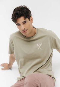 Maglietta in cotone verde oliva con scollo rotondo, caratterizzata da una grafica rosa di utensili incrociati sul petto. Texture morbida e vestibilità comoda.