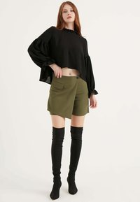 Blusa nera cropped con scollatura a volant, abbinata a pantaloni corti color verde oliva con tasche laterali e orlo asimmetrico. Stivali neri alti fino al ginocchio.