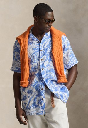 Vyras su saulės akiniais, mėlyna gėlėta trumpomis rankovėmis marškinėliais, baltais kelnėmis ir oranžiniu megztiniu, užmestu ant pečių, neutralioje fone.