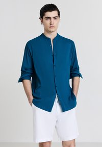 Junger Mann mit dunklen Haaren, der ein türkisfarbenes langärmliges Hemd mit Knöpfen und weiße Shorts trägt, mit seinen Händen in den Taschen vor einem schlichten Hintergrund.