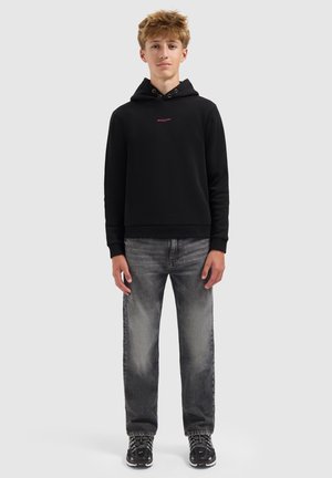 Jong persoon die staat, gekleed in een zwarte hoodie met roze tekst, vervaagde grijze spijkerbroek en zwart-witte sneakers, tegen een effen achtergrond.