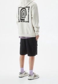Sudadera gris claro con diseño gráfico negro en la espalda, combinada con pantalones cortos cargo negros, calcetines morados y zapatillas blancas con detalles grises.