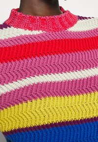 Flerfärgad stickad tröja med röd, rosa, gul och blå zick-zackmönster. Rund ribbades halsringning och textil, vävt tyg.