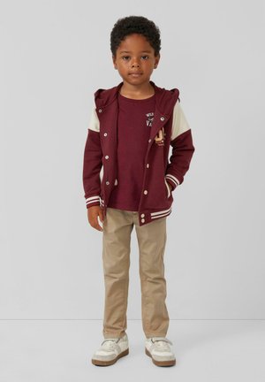 Bordeaux met rits jack met witte mouwen, beige broek en witte sneakers. Het jack heeft decoratieve knopen en een logo op het shirt eronder.