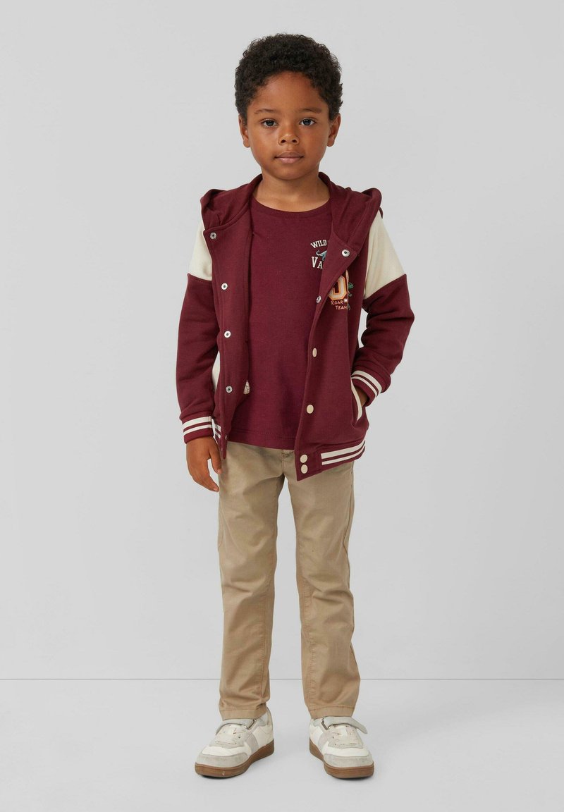 Bordeauxfarbener Zip-Hoodie mit weißen Ärmeln, beigen Hosen und weißen Turnschuhen. Der Hoodie hat dekorative Knöpfe und ein Logo auf dem darunter liegenden Shirt.