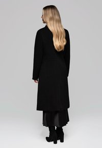 Donna bionda con capelli lunghi che indossa un lungo cappotto nero, una gonna midi nera e stivali con tacco neri, in piedi di spalle.