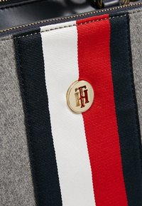 Grå stof taske med lodrette striber i rød, hvid og navy blå. En cirkulær gul logo medaljon med røde accenter er synlig.