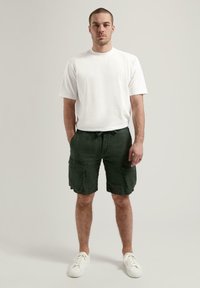 Weißes T-Shirt mit Rundhalsausschnitt, kombiniert mit dunkelgrünen Cargo-Shorts. Die Shorts haben mehrere Taschen und einen Kordelzug in der Taille. Weiße Sneakers.