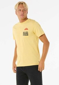 Gul bomulds t-shirt med korte ærmer, med et brystlomme-design og sort tekst, der står "RIP CURL SOAKING UP THE SUN."