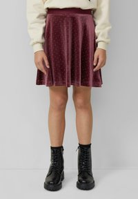 Eine Frau trägt einen weinroten Samtrock mit kleinen weißen Punkten, einen cremefarbenen Pullover, schwarze Socken und schwarze Schnürstiefel und steht an einer grauen Wand.