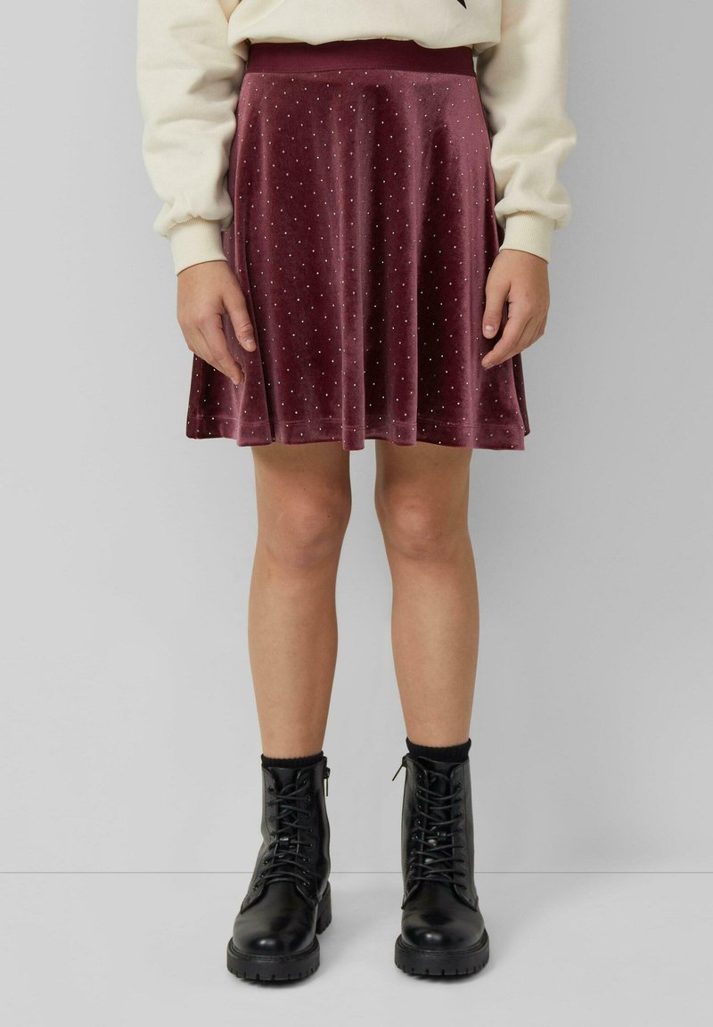 Eine Frau trägt einen weinroten Samtrock mit kleinen weißen Punkten, einen cremefarbenen Pullover, schwarze Socken und schwarze Schnürstiefel und steht an einer grauen Wand.