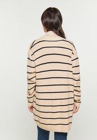 Beige pletený kardigan s černými vodorovnými pruhy, dlouhými rukávy a volným střihem. Má zakulacený spodní lem a žebrované manžety.