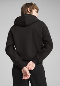 Felpa nera con cappuccio e cordino, caratterizzata da una vestibilità comoda e una texture morbida. Il modello ha le mani intrecciate dietro la schiena, mostrando la vista posteriore.