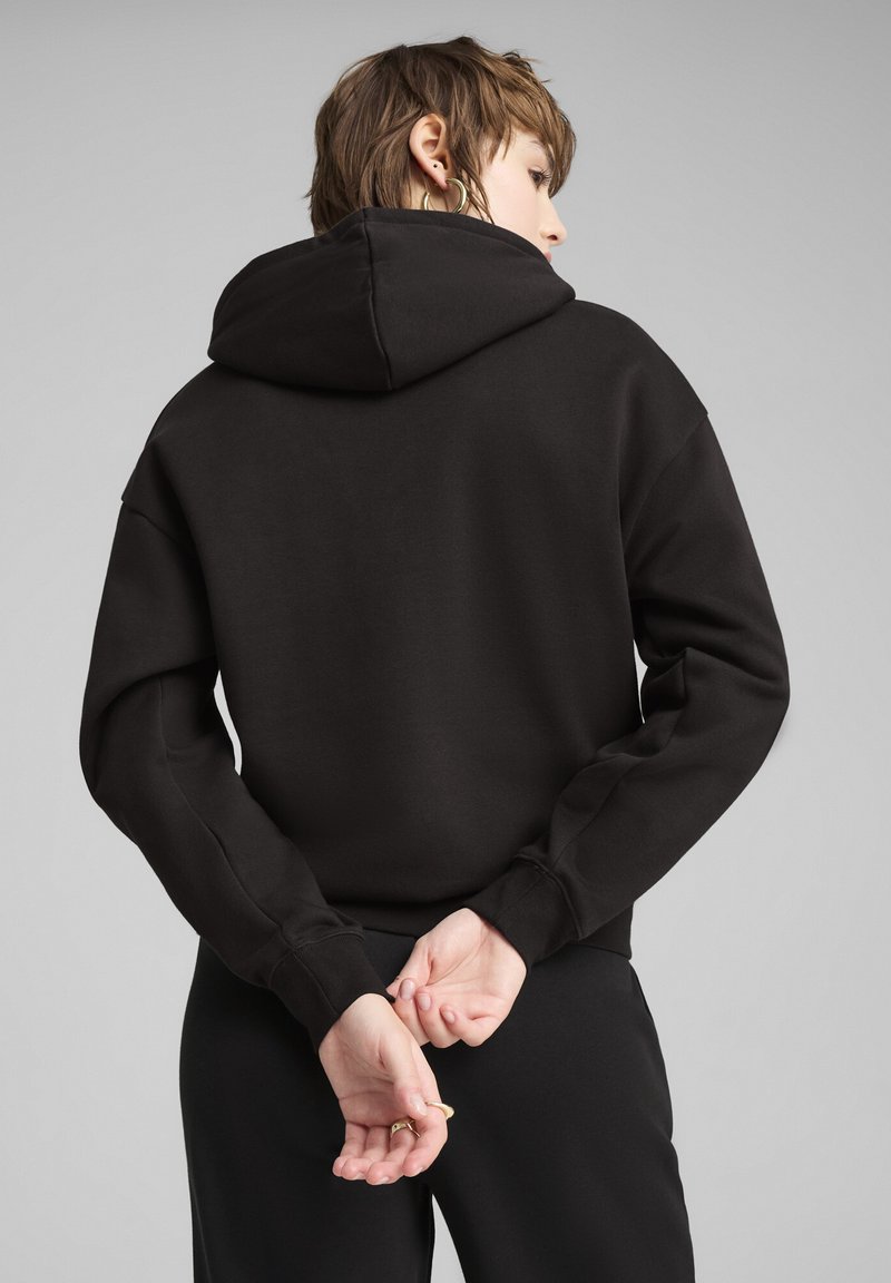 Felpa nera con cappuccio e cordino, caratterizzata da una vestibilità comoda e una texture morbida. Il modello ha le mani intrecciate dietro la schiena, mostrando la vista posteriore.