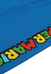 Blue fabric with a smooth texture featuring a colorful, bold graphic spelling "SUPER MARIO" in red, green, yellow, and black at the bottom edge.