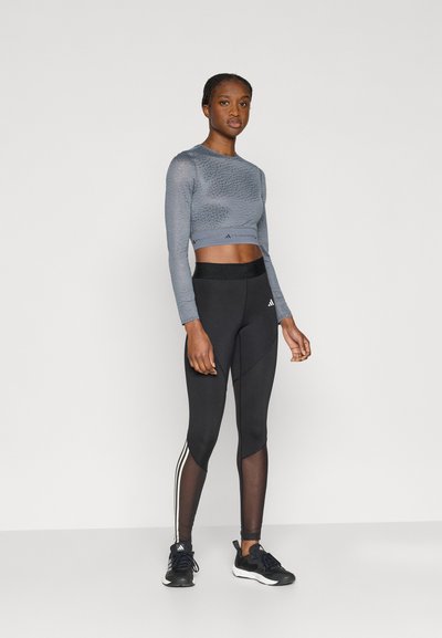 Γκρι κοντό τοπ με μακριά μανίκια και υφή, συνδυασμένο με μαύρο ψηλόμεσο κολάν που έχει λευκή ρίγα στο πλάι. Μαύρα αθλητικά παπούτσια.