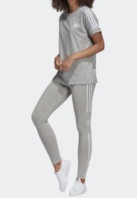 T-shirt atletico grigio con maniche arrotolate e logo bianco, abbinato a leggings grigi con strisce laterali bianche. Scarpe da ginnastica bianche completano il look.