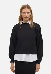 Sudadera negra con mangas voluminosas, tejido texturizado y cuello redondo, superpuesta sobre una camisa blanca con cuello, que ofrece un contraste en colores.