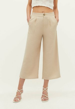 Vrouw draagt hoog getailleerde beige wijde cropped broek en witte sandalen met hak en bandjes, staand met handen in de zakken.