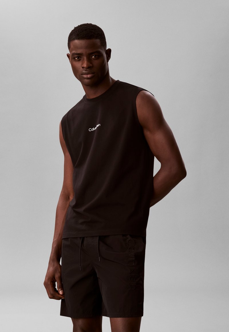 Άνδρας με μαύρο αμάνικο πουκάμισο Calvin Klein και μαύρο σορτς, στέκεται μπροστά σε μια απλή γκρίζα φόντο, κοιτάζοντας μπροστά με το ένα χέρι πίσω από την πλάτη.