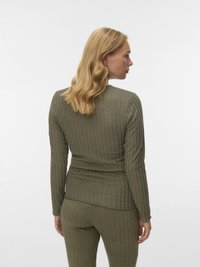 Hajú, szőke hullámos nőt látunk, aki olívazöld bordázott kötött felsőt és hozzá illő nadrágot visel, háttal a kamera felé, egy sima háttér előtt.