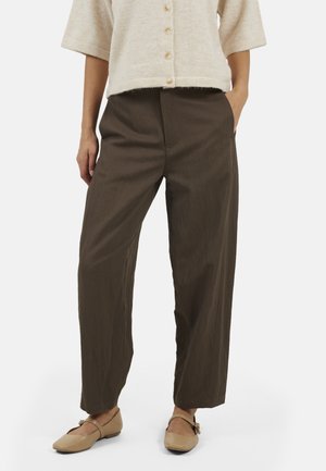 Persoon die bruine broek met brede pijpen, beige vest met knopen en beige platte schoenen met bandjes draagt, staat met één hand in de zak.