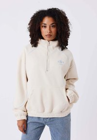 Pullover in pile di colore crema con zip a quarto, maniche raglan e tasca frontale. Presenta cuciture e dettagli del logo in azzurro chiaro.