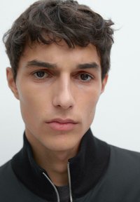 Primo piano del volto maschile con capelli corti, scuri e ricci, che indossa una giacca nera con colletto alto e cerniera, tono della pelle neutro e labbra discrete.