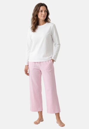 Donna in piedi scalza con una camicia bianca a maniche lunghe e pantaloni rosa chiaro a gamba larga, con una mano in tasca.