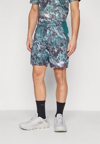 Shorts à motifs en bleu sarcelle, noir et blanc; fabriqués en tissu léger avec une taille élastique. Associés à des chaussures de course grises et des chaussettes foncées.
