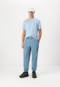Lichtblauwe gestreepte T-shirt met een geometrisch ontwerp, gecombineerd met lichtblauwe broek met elastische taille en donkere sneakers. Witte pet op het hoofd.