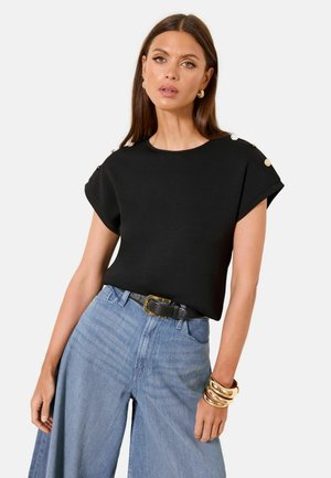 Frau mit langen dunklen Haaren, die ein schwarzes Kurzarmoberteil mit Knopfdetails, hoch taillierte, weit geschnittene blaue Jeans, goldene Creolen und Armreifen trägt.