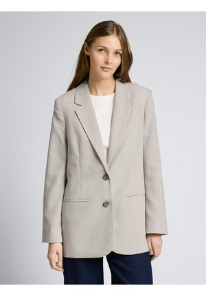 Kobieta z długimi brązowymi włosami, ubrana w jasnoszary, oversize'owy żakiet, biały top i ciemne spodnie, stojąca na tle gładkiego, szarego tła.