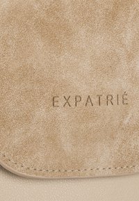 Borsa in finta pelle beige con finitura testurizzata. Presenta un logo in rilievo con la scritta "EXPATRIÉ" sulla patta. Bordo liscio e dettagli di cucitura.