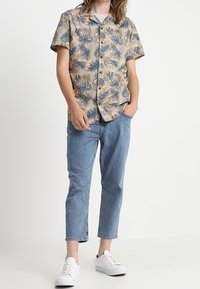 Short-sleeve, beige button-up shirt with a blue and brown leaf pattern, paired with light blue jeans and white sneakers.