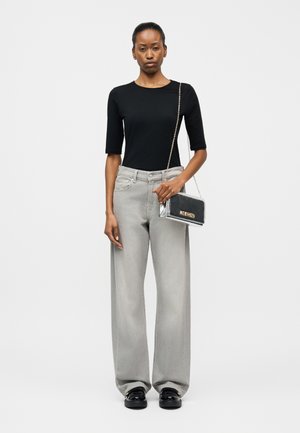 Frau trägt ein schwarzes Oberteil mit Ellbogenärmeln, hellgraue Jeans mit weitem Bein, schwarze Lackschuhe und hält eine kleine silberne Metallic-Tasche mit Kettenriemen.