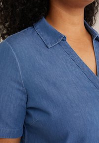 Chemise en denim bleu avec un col, des manches courtes, et une coupe décontractée avec une texture subtile et des détails de couture.