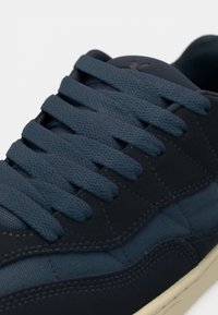 Sneaker blu scuro con lacci spessi e testurizzati, pannelli in suede e tessuto, e suola beige, mostrato in una vista ravvicinata.