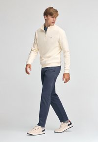 Sudadera cremosa con cremallera, pantalones azul marino y zapatillas beigas. La sudadera tiene un logotipo sutil; los pantalones tienen un corte recto.