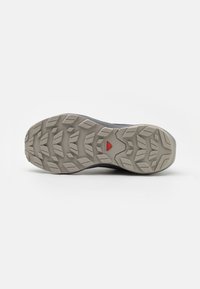 Semelle d'une chaussure de randonnée grise avec des motifs de semelle profonds et robustes et un logo triangulaire rouge au centre.