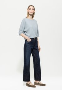 Helles blaues Strickpullover mit lässigen Ärmeln, kombiniert mit dunkelblauen Jeans in gerader Schnittform und grünen Ballerinas mit einem Riemendetail.