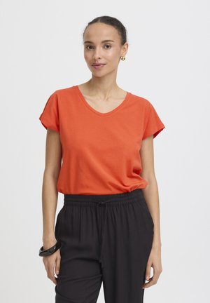 Nő összefogott hajjal, arany karikafülbevalókat viselve, narancssárga, rövid ujjú, V-nyakú pólóban, fekete zsinóros nadrágban és fekete karkötővel.