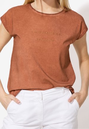 Frau trägt ein rostfarbenes kurzärmeliges Oberteil mit dem Text „I'M WALKING ON SUNSHINE“ und weiße Hose, Hände in den Taschen.
