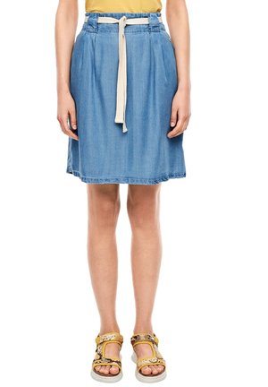 Person iført en knælængde blå denim nederdel med et hvidt bælte, gul top og gule mønstrede sandaler, stående mod en hvid baggrund.