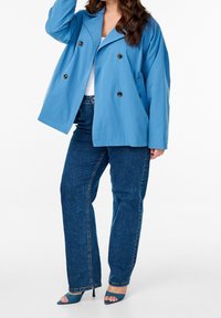 Žena ve dvojřadém modrém saku, tmavě modrých džínech s rovnými nohavicemi a modrých otevřených botách na podpatku, pózující na bílém pozadí.