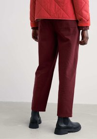 Osoba w ciemnoczerwonych spodniach, czarnych botkach za kostkę i czerwonej pikowanej kurtce stoi na jasnoszarym podłodze w obrębie gładkiej ściany.