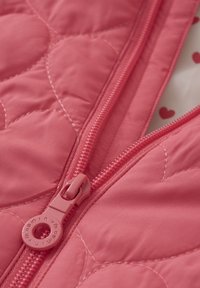 Trapetsärmad rosa väst med en central dragkedja, rund dragkedjeknopp och hjärtformade sömmönster på tyget.
