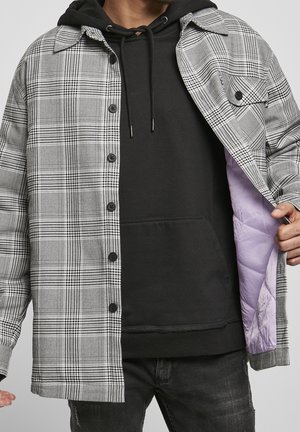 Mann trägt schwarzen Kapuzenpullover und graue karierte Hemdjacke mit lavendelfarbenem Steppmuster, öffnet die Jacke, um das Innenfutter zu zeigen.