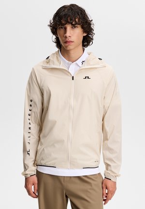 Young man with curly hair wearing a beige zip-up jacket with black logo and writing on the left sleeve, white polo, and brown pants.